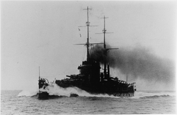 A large battleship steaming through the water at high speeds. Water breaks against the bow of the ship while smoke billows from the funnels.