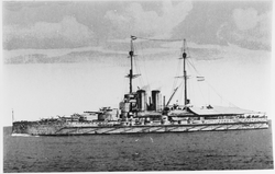 A large battleship steams through the water. Small amounts of smoke can be seen coming from the ship's funnels.