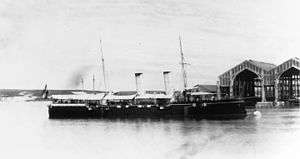 A large warship with a black hull moored in harbor, with canvas awnings covering the deck