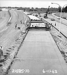 A black and white photograph of a concrete road in the middle of paving. Piles of dirt, forming a ramp, can be seen in the background.