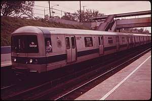 A Staten Island Railway train composed R44 subway cars on the Staten Island Railway. This image, taken in 1973, shows the cars with a since-removed blue stripe toward the bottom of the car body. The train is arriving at a platform to the left; the photo is taken from another platform to the right and in the foreground. The station is in a right-of-way below street level, and a covered footbridge connecting the two platforms is located to the right.