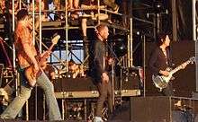 A rock band, the Stone Temple Pilots, performing onstage. From left to right, a bass guitarist, singer and guitarist are shown.