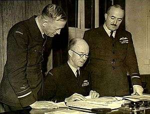 Three men in military uniforms, two standing and one seated, looking at papers on a desk