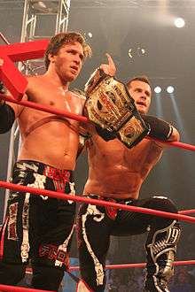 Two male wrestlers standing in a ring holding championship belts wearing black wrestling gear.