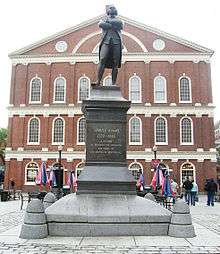 A statue on a pedestal of a man standing with his arms crossed. An inscription on the pedestal reads, "Samuel Adams, 1722–1803. A Patriot. He organized the Revolution and signed the Declaration of Independence." Behind the statue is a three-story brick building with many windows.