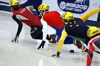A tight group of four skaters leaning inwards as they make a turn.  The four skaters are wearing yellow helmets and suits that the display flag colors of their respective countries.  The skaters have their left hands touching the ice for balance as they accelerate around the turn.