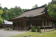 Color photo of a one-storey wooden building of a Buddhist temple with a thatched roof.