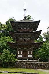 A brown wooden pagoda on a stone base. In the background: trees with green foliage.