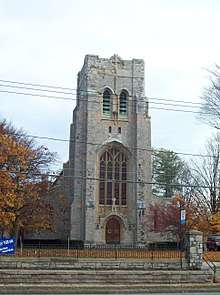 A stone church tower with a level top, gently arched pointed windows and a small wooden door in the base. In front of it are telephone lines and a set of steps.