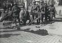 Seven soldiers of the German Army are stationed at a street corner in Helsinki after the surrender of the Red Guard headquarters Smolna. One of them is on his knee while two are relaxing against a railing or on a chair. MG 08, a heavy machine gun, rests in front of them.