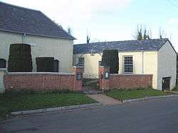 Two single-storey cream buildings with slate roofs at an angle to each other. On the left is part of the chapel; on the right is the assembly hall with two sash windows.  In front is a brick wall and gateposts; above the gate is an overthrow with a lantern.