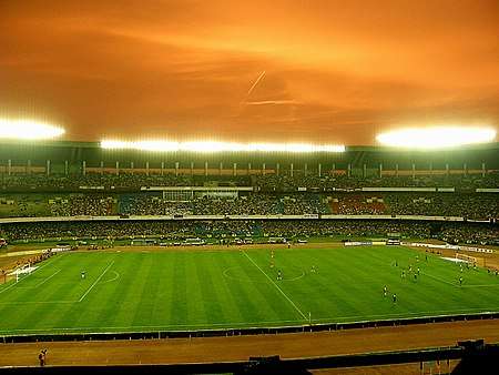 A large football stadium which is roughly 75% full. There are footballers on the pitch, most of whom are on the right side of the stadium. The team on the left, East Bengal are wearing red while Bayern Munich, right, are wearing black. The floodlights are on and the sky appears to be orange.