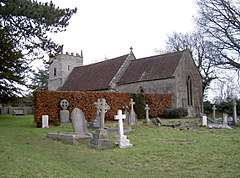 Grey stone building with red tiled roof, partially obscured by a hedge. A square tower is at the far end. The foreground includes several crosses and gravestones.