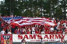 Soccer fans, dressed in red, cheer in bleachers as they hold a large American flag over themselves at a soccer match.