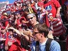 A number of American fans, dressed in red, stand in bleachers holding United States paraphernalia.