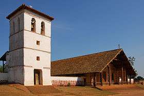 A church and stone bell-tower in three-quarter view. The whitish facade of the church is decorated with motives painted in orange. A wooden cross is positioned at the top of the roof.