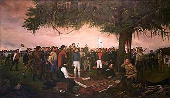 A group of men are gathered under a large tree.  One man lays on the ground under the trees, with his bare foot exposed.