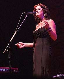 Middle-aged woman with medium-length brown hair wearing a low-cut evening dress while singing into a microphone with her eyes closed.