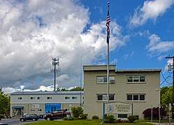 A buff-colored two-story flat-roofed building under a blue sky with clouds. To the left rear is a garage and a cell-phone tower. In front is a flagpole and a large wooden sign reading "Col. Roger B.C. Donlon Town Hall".