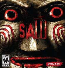 The box art features the face of an antique-looking Billy doll. The face is gold in color and is shiny. It has red eyes and red lips. The word "Saw" in red letters is in the center.