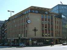 A 1950s or 1960s six-story building, predominantly made of yellow brick with red brick used around the windows. Other ornamentation include a large round black and white clock, a large black cross symbol, and "Scientology Kirche Hamburg e.V." lettering.