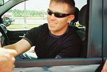 A man in his thirties with a head full of hair. He is wearing sunglasses and a black T-shirt and is shaking hands with another person.