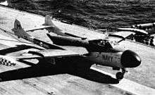 A jet aircraft with a twin tail arrangement sitting on the flight deck of an aircraft carriers. Cables trailing from underneath the aircraft indicate that it has just landed.
