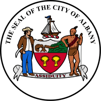 A black circle with white fill is shown with the coat of arms at center. Above the coat of arms, following an arced path, are the letters "The Seal of the City of Albany"