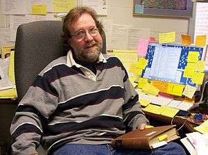 Professor Sean B Carroll sitting at his desk, holding a book, with lots of post-it notes glued everywhere.