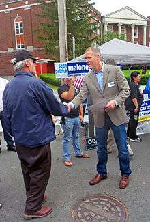 A man wearing a jacket, shirt, dark jeans and loafers, leaning forward and shaking hands with an older man wearing a black baseball hat and blue windbreaker.