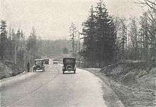 A black-and-white photograph of a paved road cutting through a wilderness-like scene. Four automobiles can be seen on the road, traveling in both directions, along with a man waiting on the shoulder.