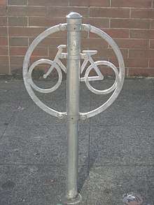 A ring with a stylized bicycle inside, with riveted to a post (running through the ring), all of galvanized metal. It is set in a concrete slab in front of a brick wall.