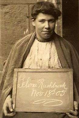 short-haired woman in prison smock, holding a blackboard with "Selina Rushbrook Nov 18 05" written on it