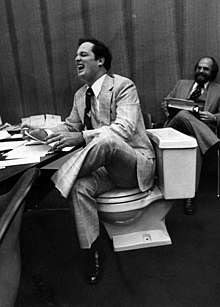 A laughing man wearing a suit sits on a toilet. In front of him is a desk with papers.