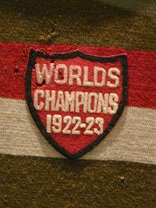 A red shield-shaped cloth crest with the inscription "Worlds Champions 1922–23" sewn onto a gold red and white striped sweater