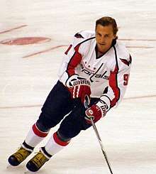 A hockey player in a blue, red and white uniform looks into the distance as he makes a turn while skating.