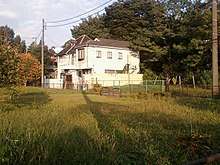 A white, two-story house surrounded by trees with a large, empty field of grass in the foreground