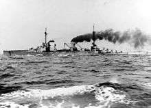 A large gray battlecruiser steams through choppy seas, thick black smoke pours from its rear smoke stack.