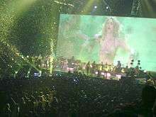 An image of a woman dancing is being projected onto a large screen. There is green lighting in the entire arena and large amounts of confetti are floating in the air.
