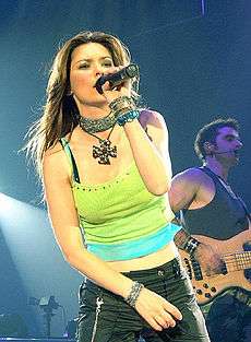 A woman with long brown hair wearing a colourful tank top, dark pants and a large necklace, singing into a microphone