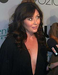 A woman with shoulder-length black hair wearing a black dress with a plunging neckline speaks to a reporter.