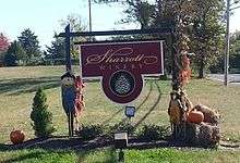 A maroon-colored sign post decorated with pumpkins, scarecrows, and bales of hay, with ground-based floodlights on both sides of the sign.