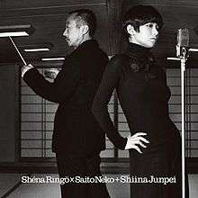 A man turned to his right in a suit holds a conductor's baton. Next to him stands a woman with short hair, standing in front of a vintage microphone stand. The pair stand in a tatami room with shoji.