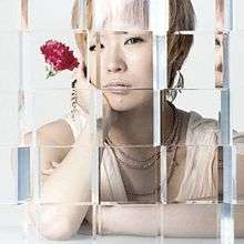 The top half of a woman sitting down in a white dress and decorative jewellery, with short, blonde hair. She is holding a red carnation. Her image is distorted and refracted in places, as it is shown through several glass cubes.