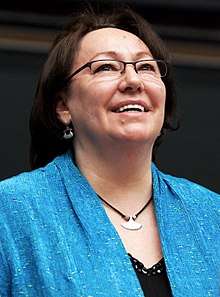 A colour photo of Sheila Watt-Cloutier, looking up and smiling, while giving a lecture at York University. She is a wearing a blue jacket over a black shirt and has an ulu necklace.