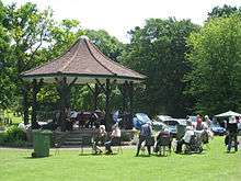 Grassy area with people sitting in chairs in front of a bandstand with tiled roof.