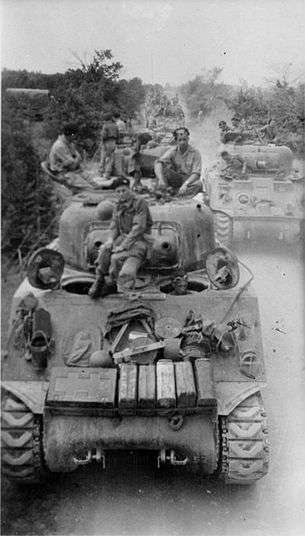 tanks with several men sitting on its turret advancing along road towards camera