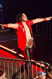 Shinsuke Nakamura, a black-haired Japanese man wearing a red vest, black pants and the white-and-gold IWGP Intercontinental Championship, struts down the entrance ramp.
