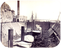 A photograph with two steamships resting in a dewatered drydock with a building housing the engine for operating the lock's gates in the background