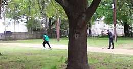 Video screenshot of Scott in a turqoise shirt and black pants running away to the left, many meters away, with Slager in uniform pointing his gun at Scott in the right of the screen